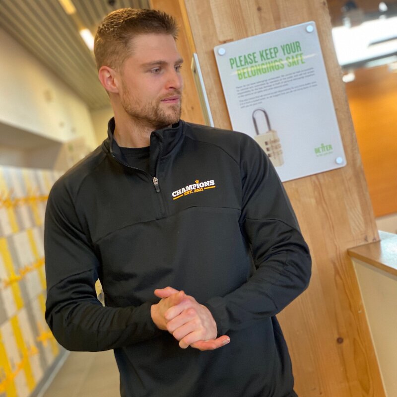 A male personal trainer in a gym changing room sporting a black customised quarter-zip sweatshirt.